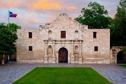 The front of The Alamo with a Texas flag, trees and sunset clouds.