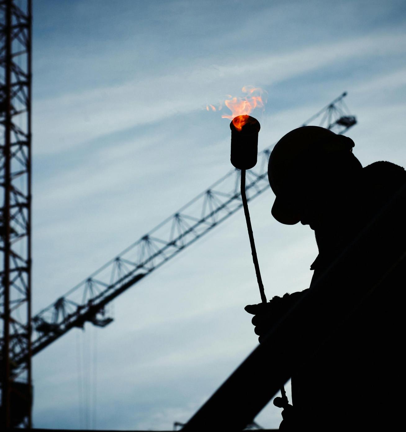 Silhouette of a construction worker holding a blowtorch with a crane and blue sky in the background.