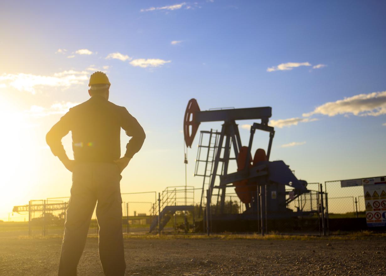 Silhouette of male worker with hardhat overlooking a pumpjack with bright sun in the background